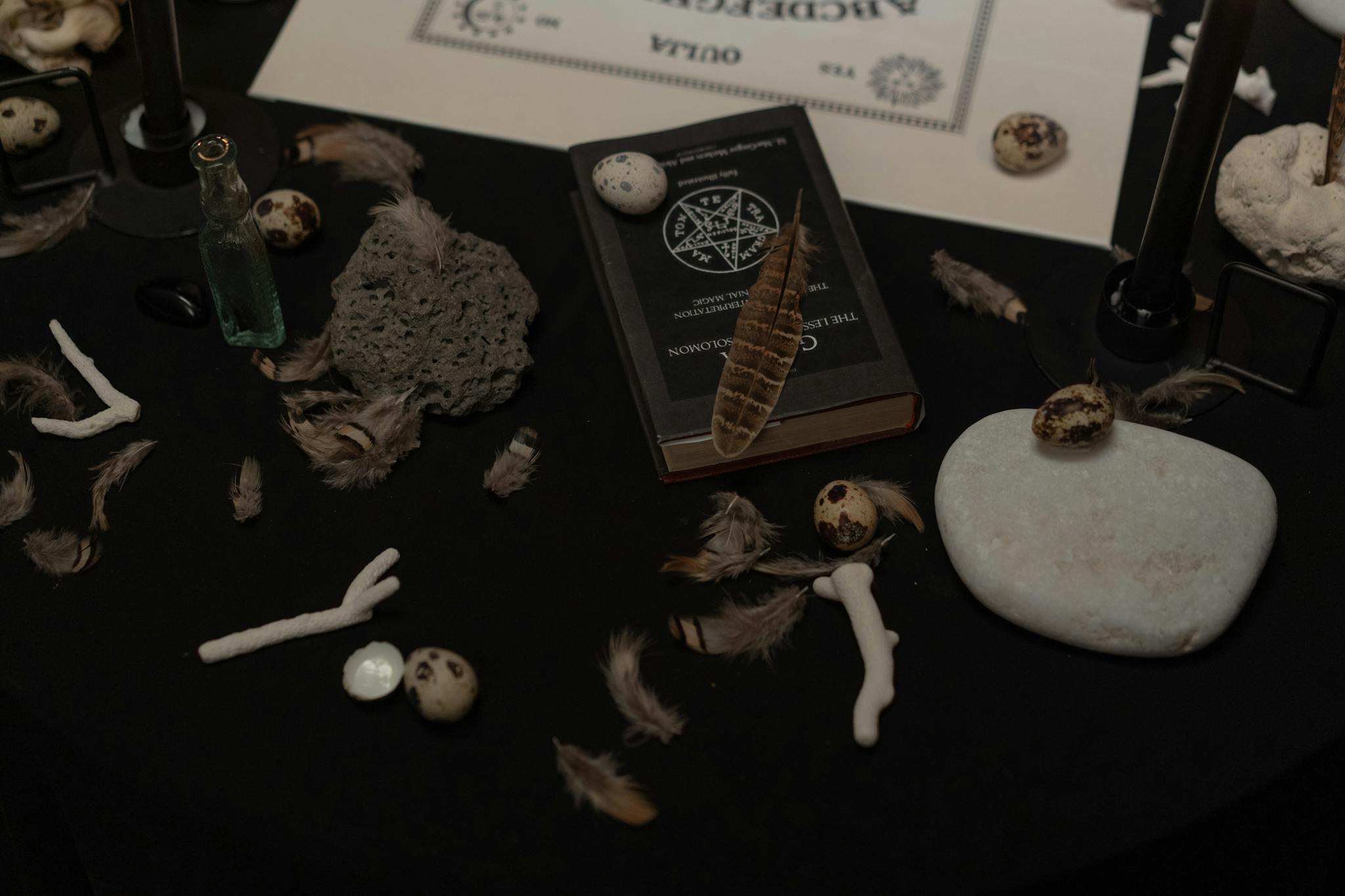 A dark, mysterious scene with a book, feathers, stones, and ritualistic items.